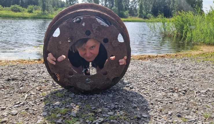 Person looking through a large circular metal object by the water.