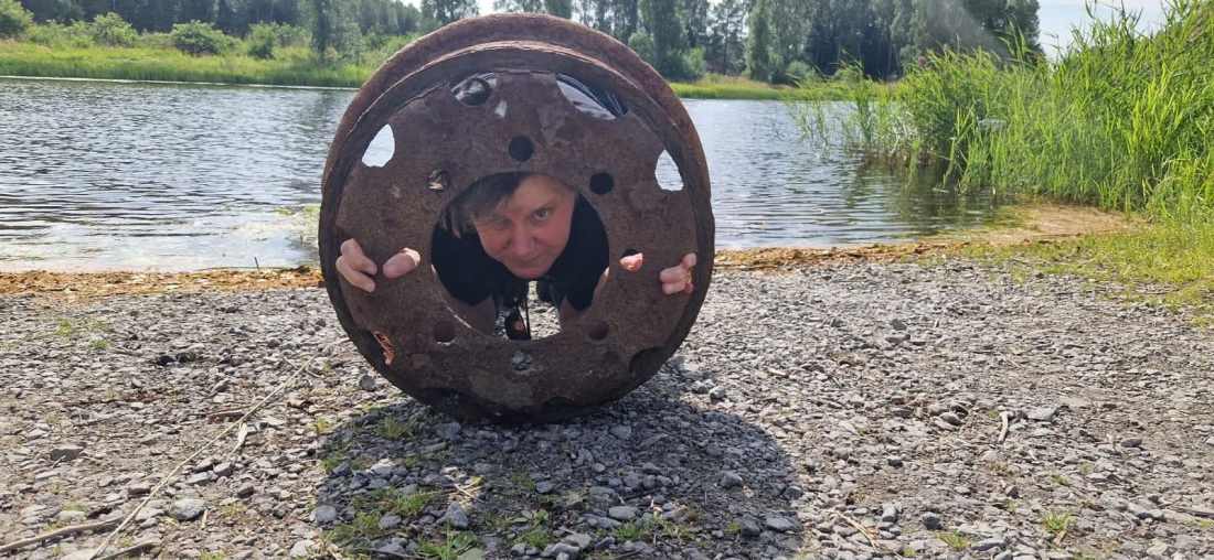 Person looking through a large circular metal object by the water.